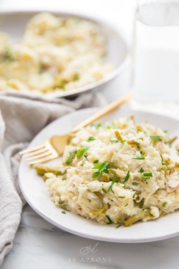 Quick creamy Whole30 cauliflower risotto with chicken and asparagus on a white plate with a gold fork, grey linen napkin next to the plate, and skillet with more risotto in the background