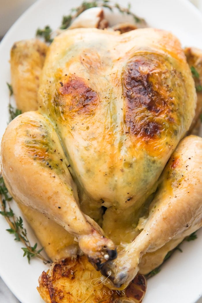 A whole chicken with golden skin on a white plate with fresh thyme and roasted garlic