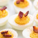 Deviled egg with a couple pieces of bacon on top surrounded by other eggs