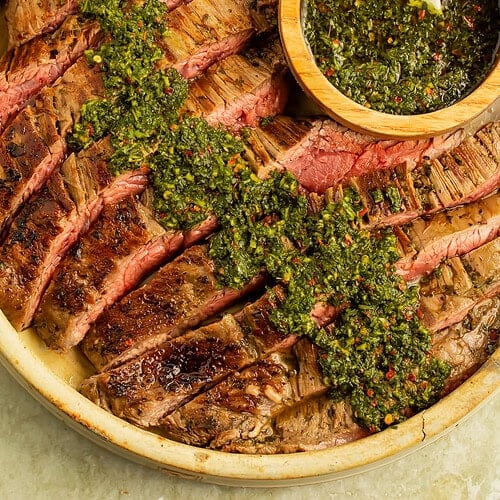 Plate of flank steak with a wooden bowl of chimichurri sauce, which has been spooned over the steak.