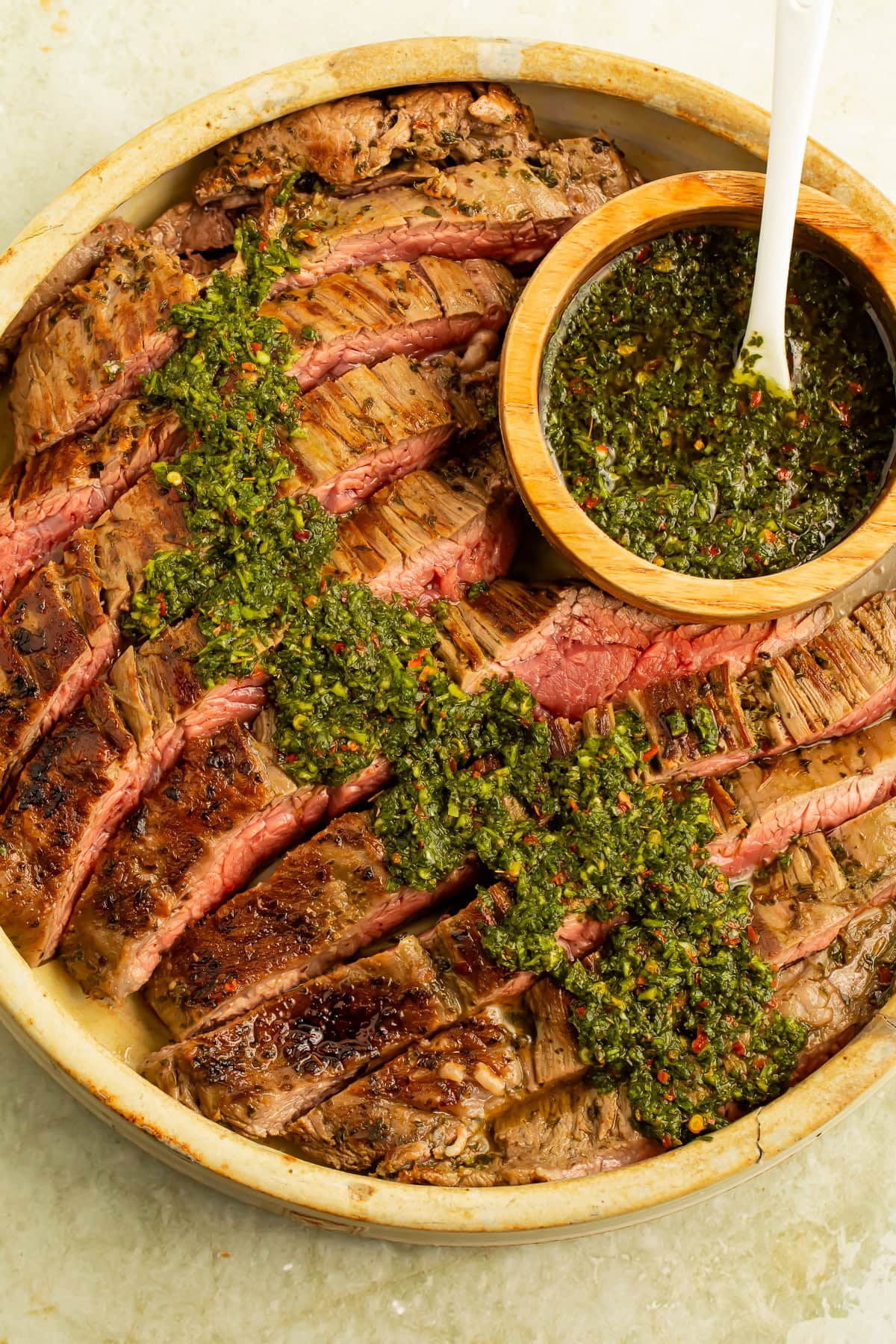 Plate of flank steak with a wooden bowl of chimichurri sauce, which has been spooned over the steak.