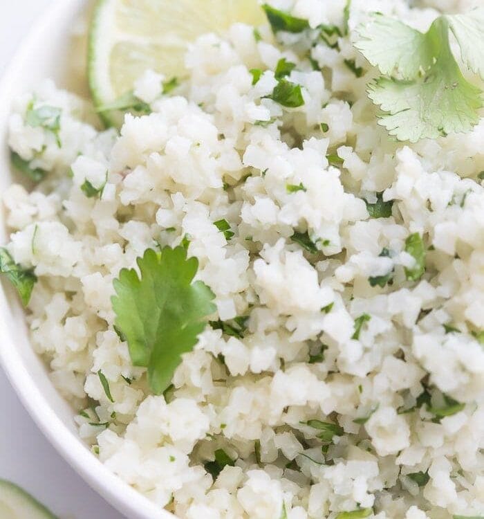 A white bowl of cilantro lime cauliflower rice with cilantro and lime garnishes
