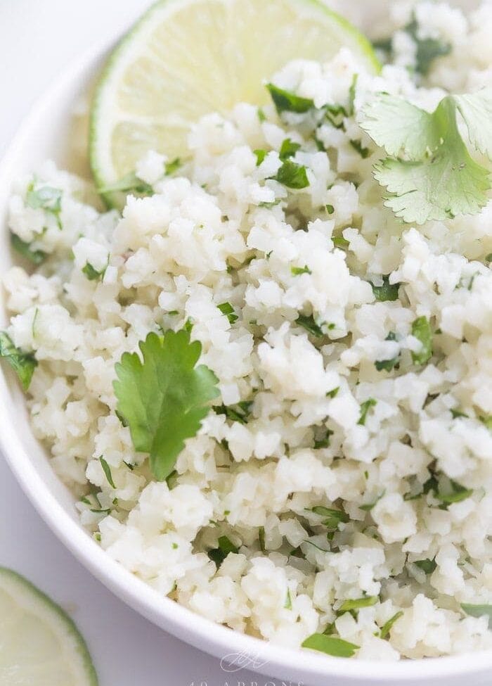 A white bowl of cilantro lime cauliflower rice with cilantro and lime garnishes