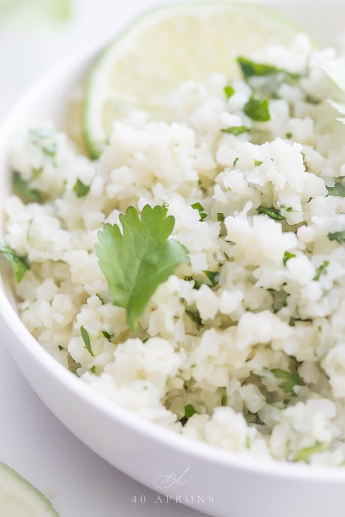 A white bowl of cilantro lime cauliflower rice with cilantro and lime garnishes.