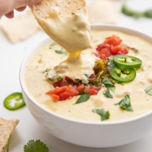 Hand dipping a tortilla chip into a bowl of vegan queso topped with tomatoes and jalapenos