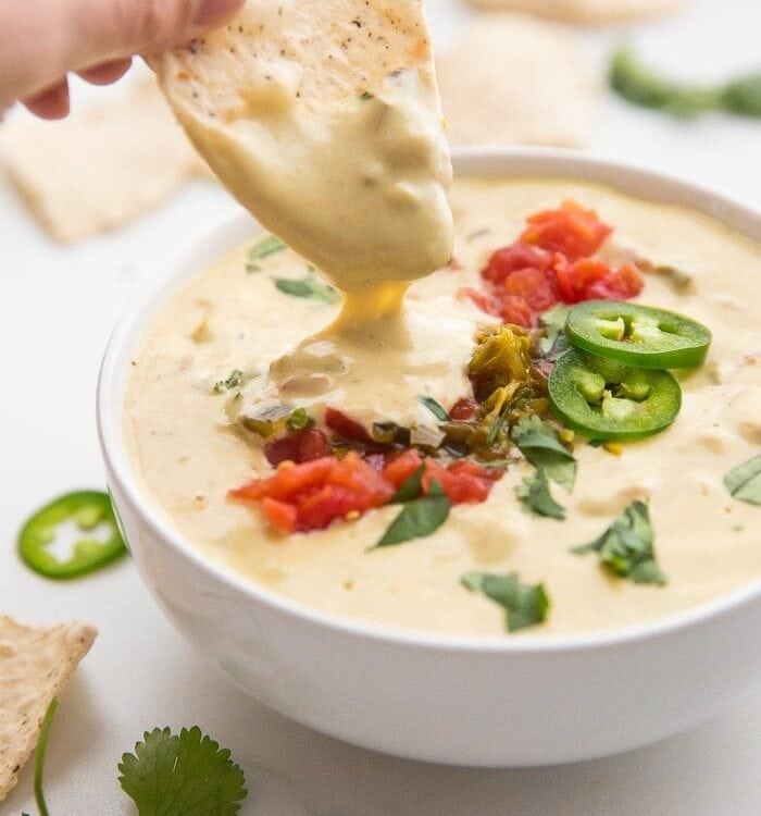 Hand dip a tortilla into a bowl of vegan queso topped with tomatoes and jalapenos