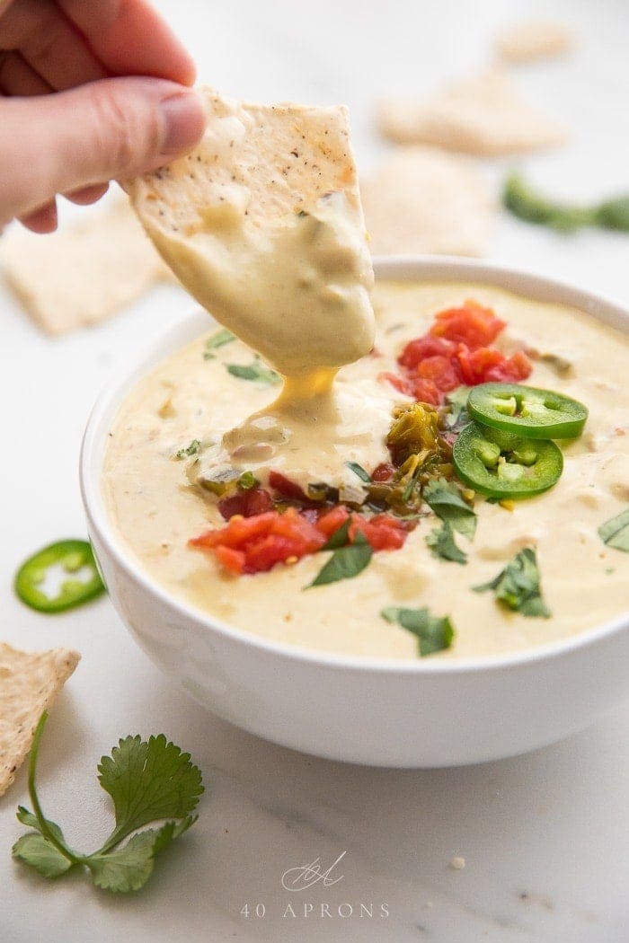 Hand dipping a tortilla chip into a bowl of vegan queso topped with tomatoes and jalapenos