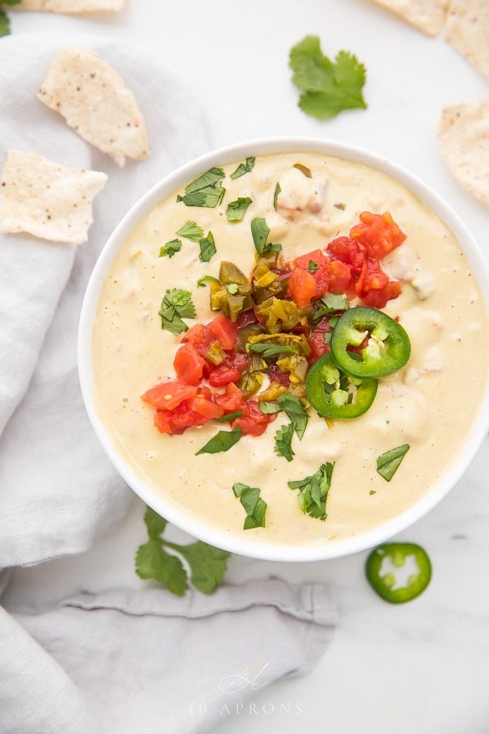 Zoomed out photo of vegan queso topped with tomatoes and jalapenos surrounded by cilantro and tortilla chips