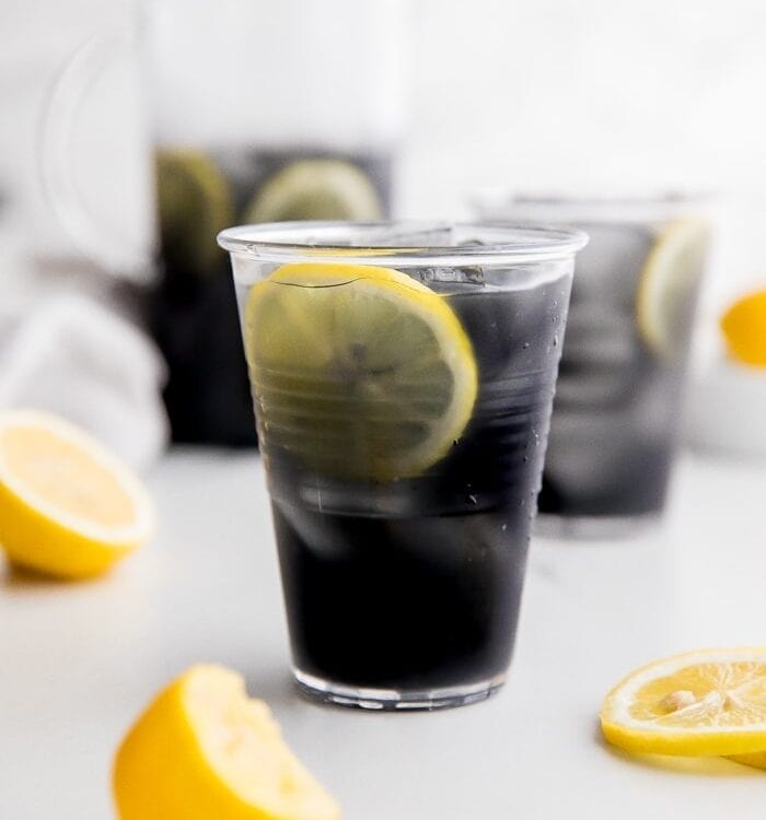 A glass of black charcoal lemonade with lemon slice, with glass and pitcher in background