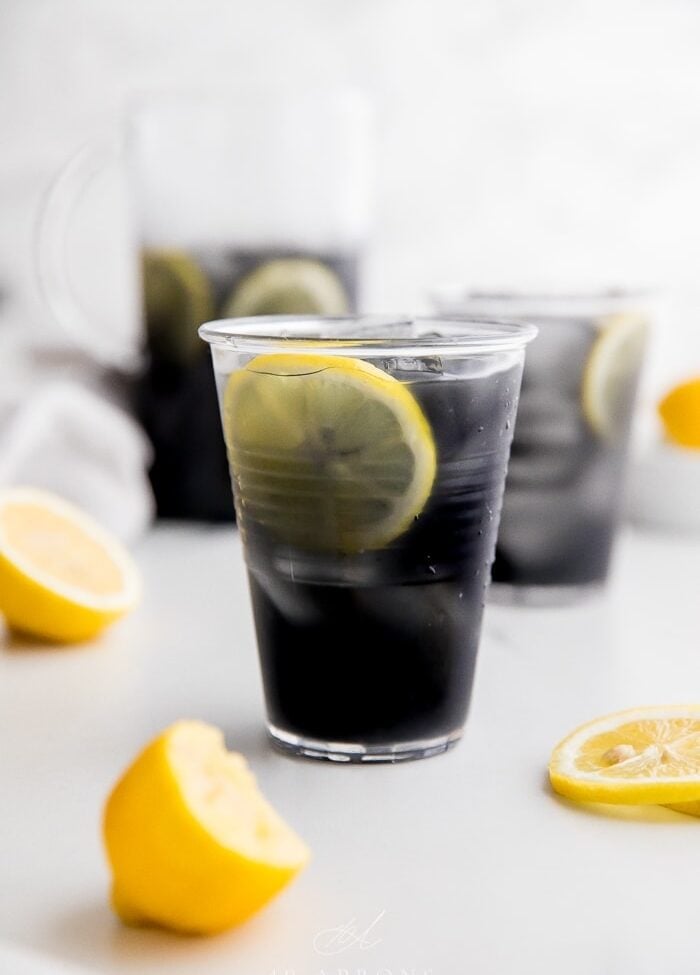 A glass of black charcoal lemonade with lemon slice, with glass and pitcher in background