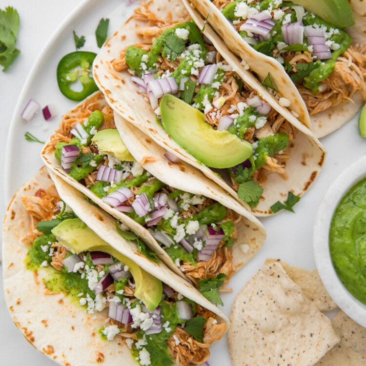 4 Crockpot chicken tacos topped with avocado, cilantro, queso fresco, and red onion lined up in a row on a platter.