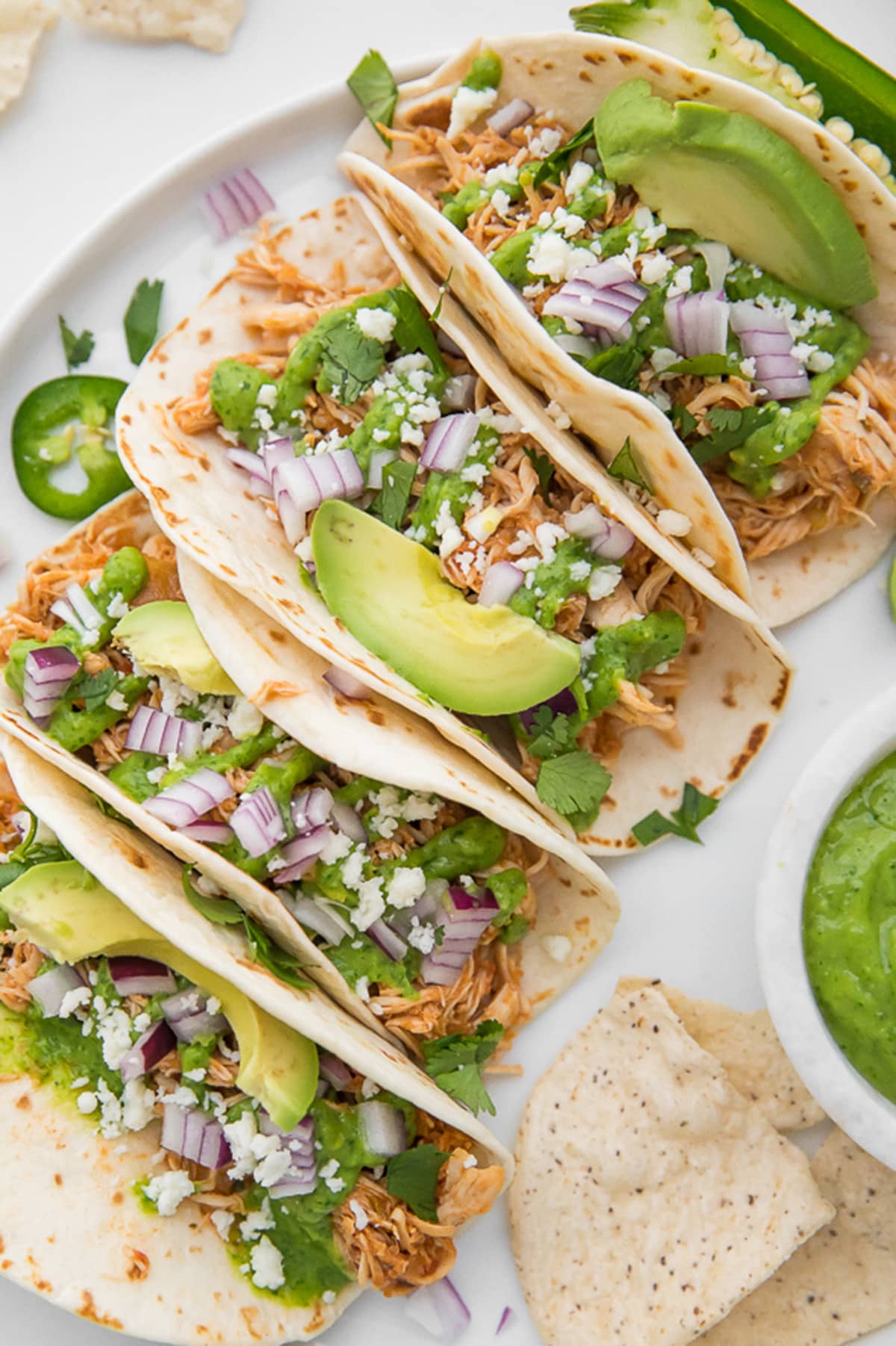 4 Crockpot chicken tacos topped with avocado, cilantro, queso fresco, and red onion lined up in a row on a platter.