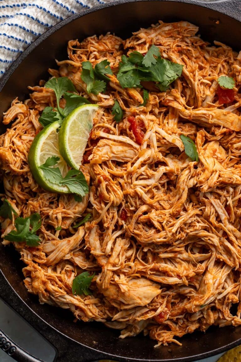 Slow cooker shredded Mexican chicken in a large cast-iron skillet, garnished with lime wedges and cilantro.