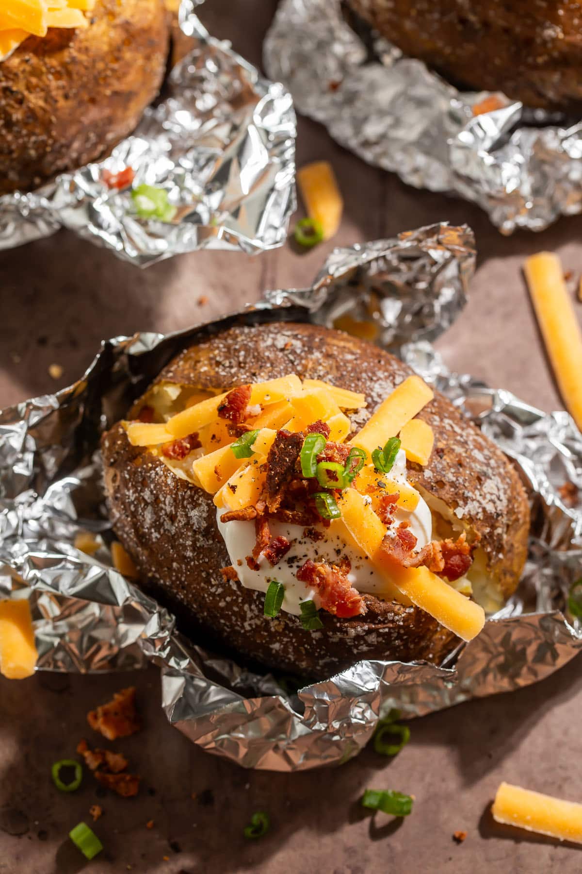 Foil-wrapped baked potatoes, cooked on a grill, split open and topped with shredded cheddar, a dollop of sour cream, and chopped green onions.
