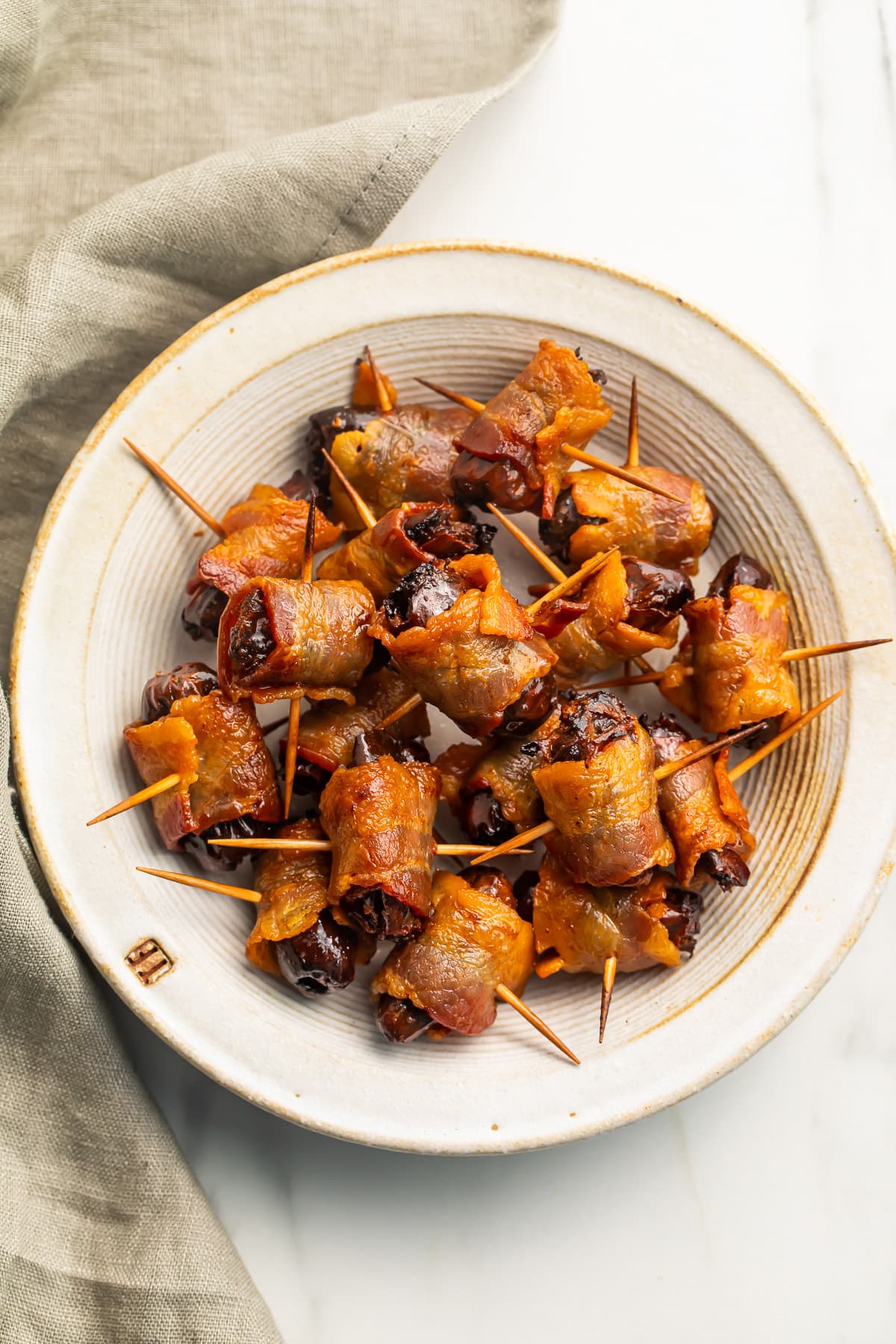 Several bacon-wrapped dates piled in a large vintage-looking bowl next to a cloth napkin.