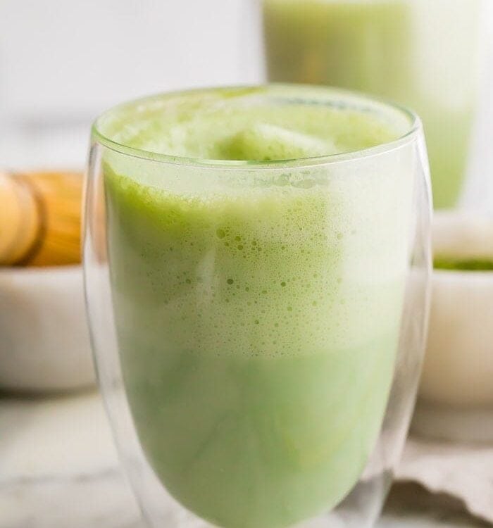 two matcha lattes in glass cups with a bamboo whisk to the side