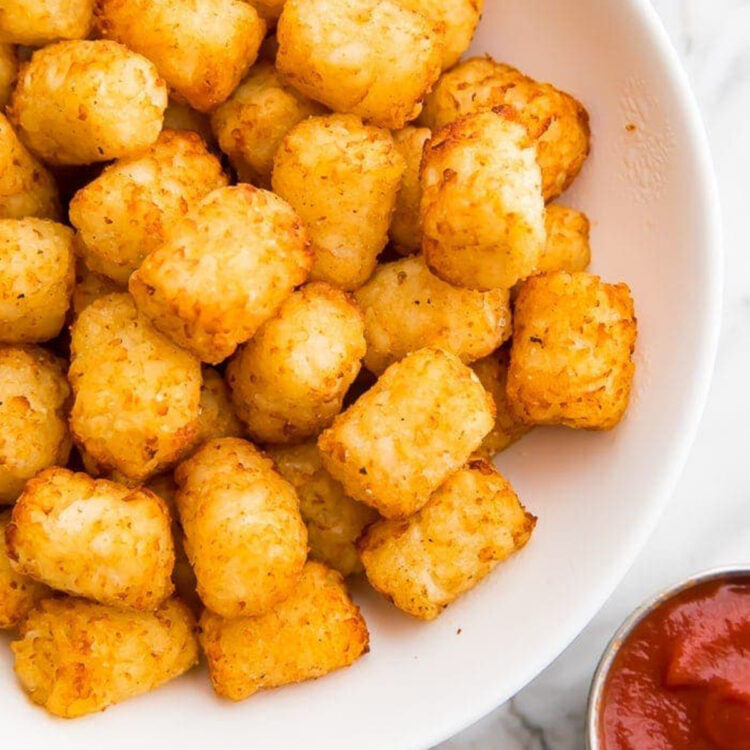 Crispy, golden air fryer tater tots in a large white serving bowl next to a small ramekin of bright red ketchup.