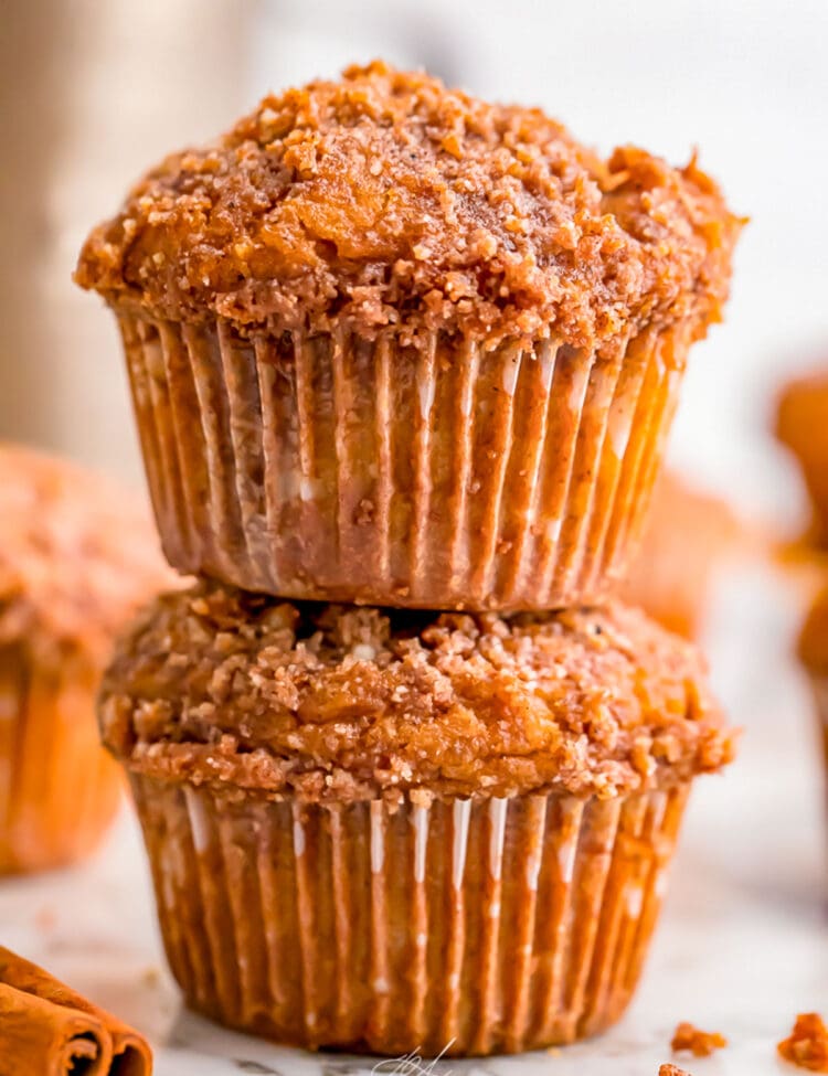 Two pumpkin muffins with a spiced crumb topping stacked on top of each other, with other muffins out of focus in the background.