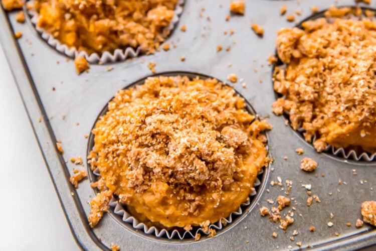 Unbaked pumpkin muffin batter in a muffin tin cavity lined with parchment paper liner, topped with a spiced crumb topping.