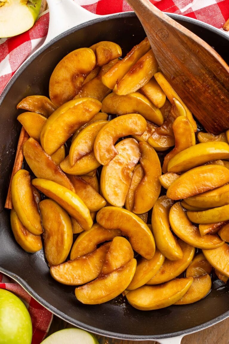 A large cast iron skillet of fried apple slices coated in brown sugar cinnamon butter resting on a table with a red and white checkerboard napkin.