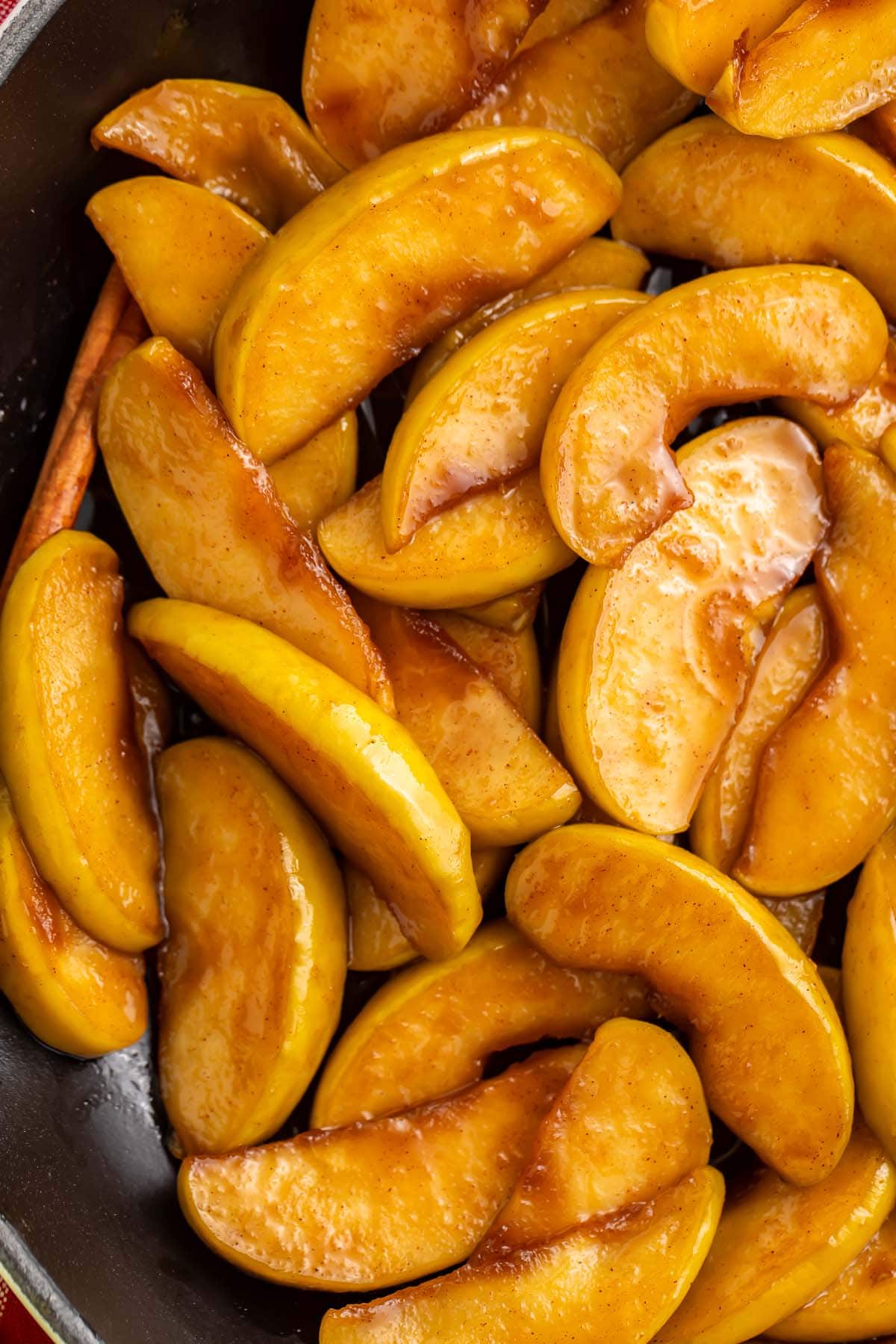 A large cast iron skillet of fried apple slices coated in brown sugar cinnamon butter resting on a table with a red and white checkerboard napkin.