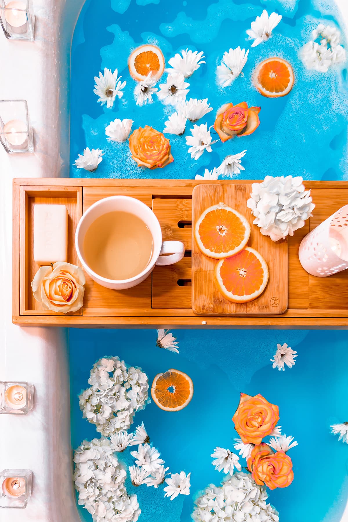 Overhead view of a bathtub filled with bright blue water and orange slices with white flowers and a wooden tray holding a cup of coffee.