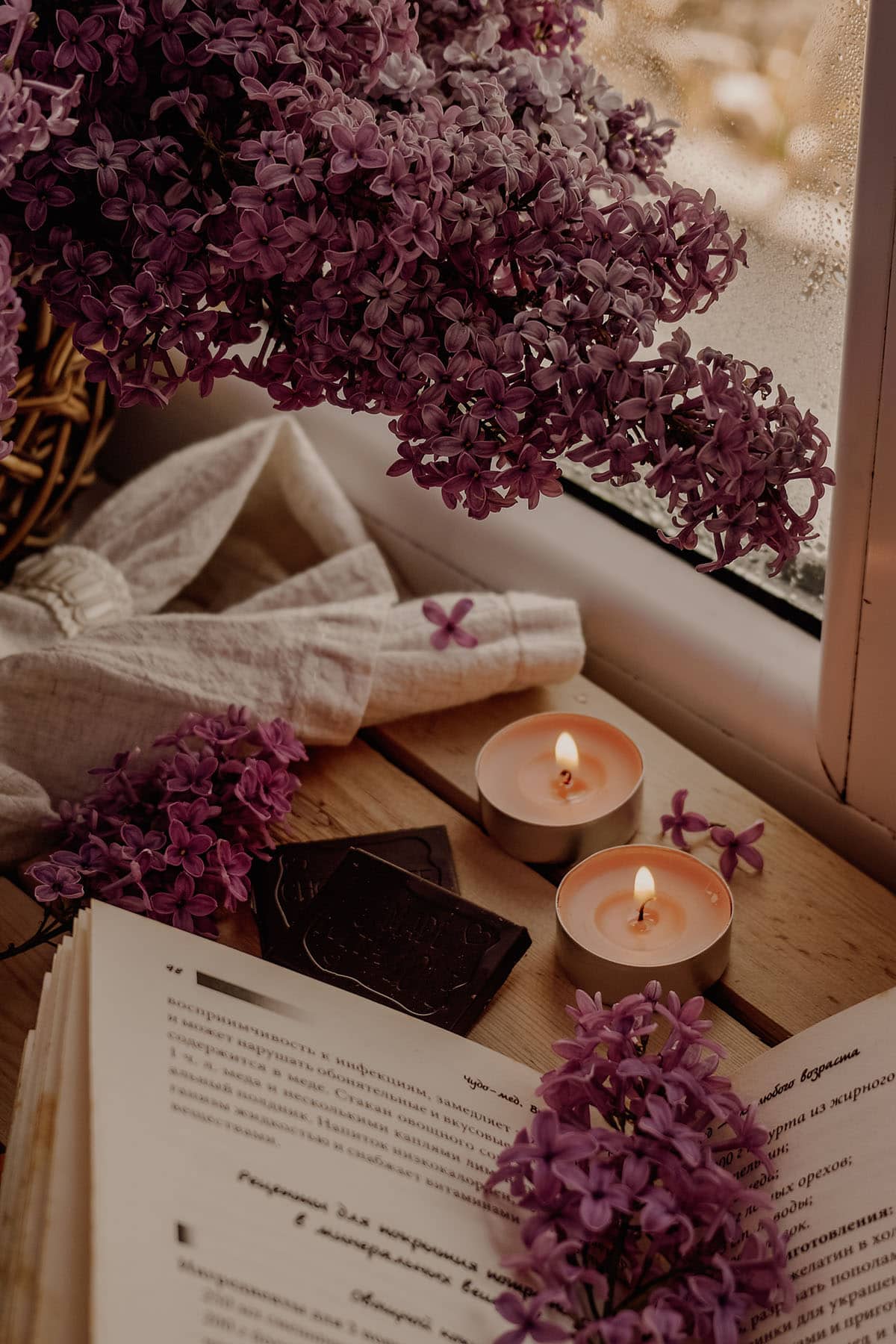 Image of lavendar flowers, two lit candles, and an open book next to a window on a peaceful morning.