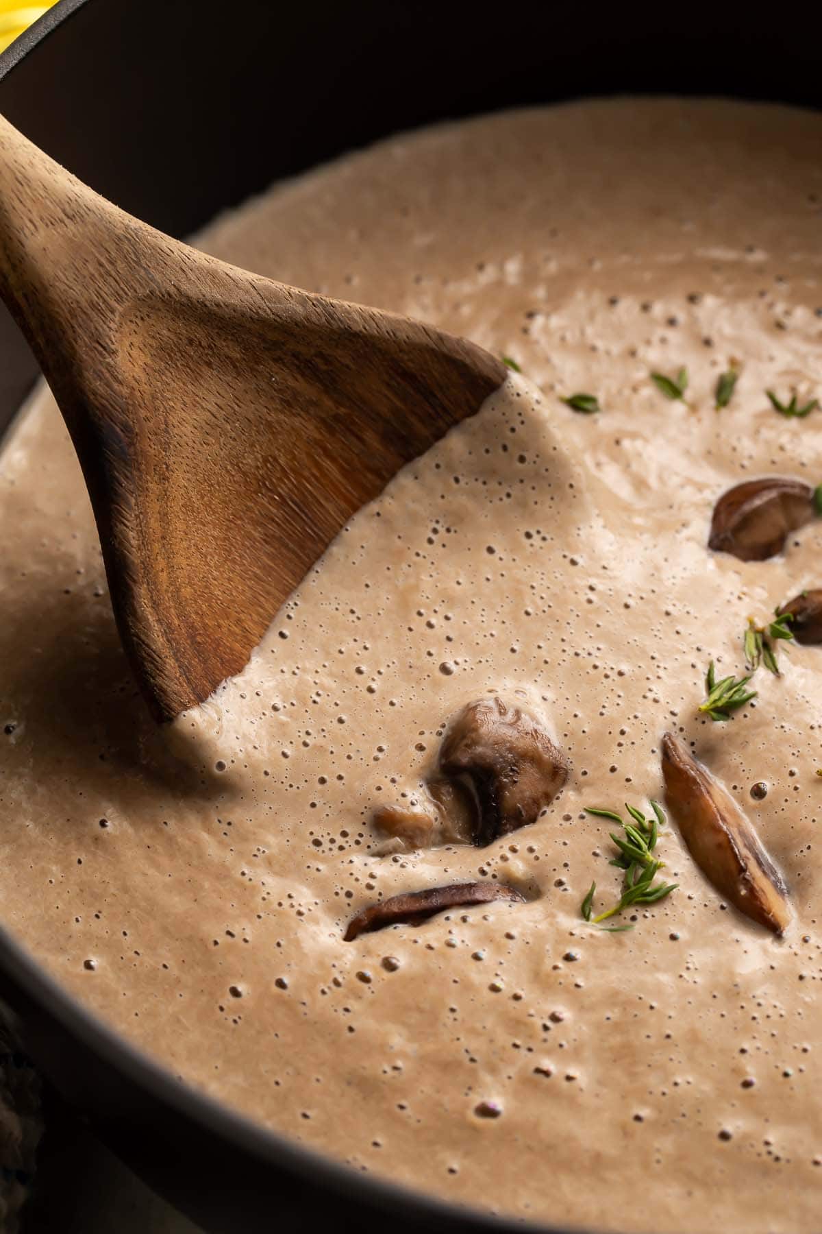 A close-up photo of a large wooden spoon stirring a creamy, thick cream of mushroom soup in a large black pot.