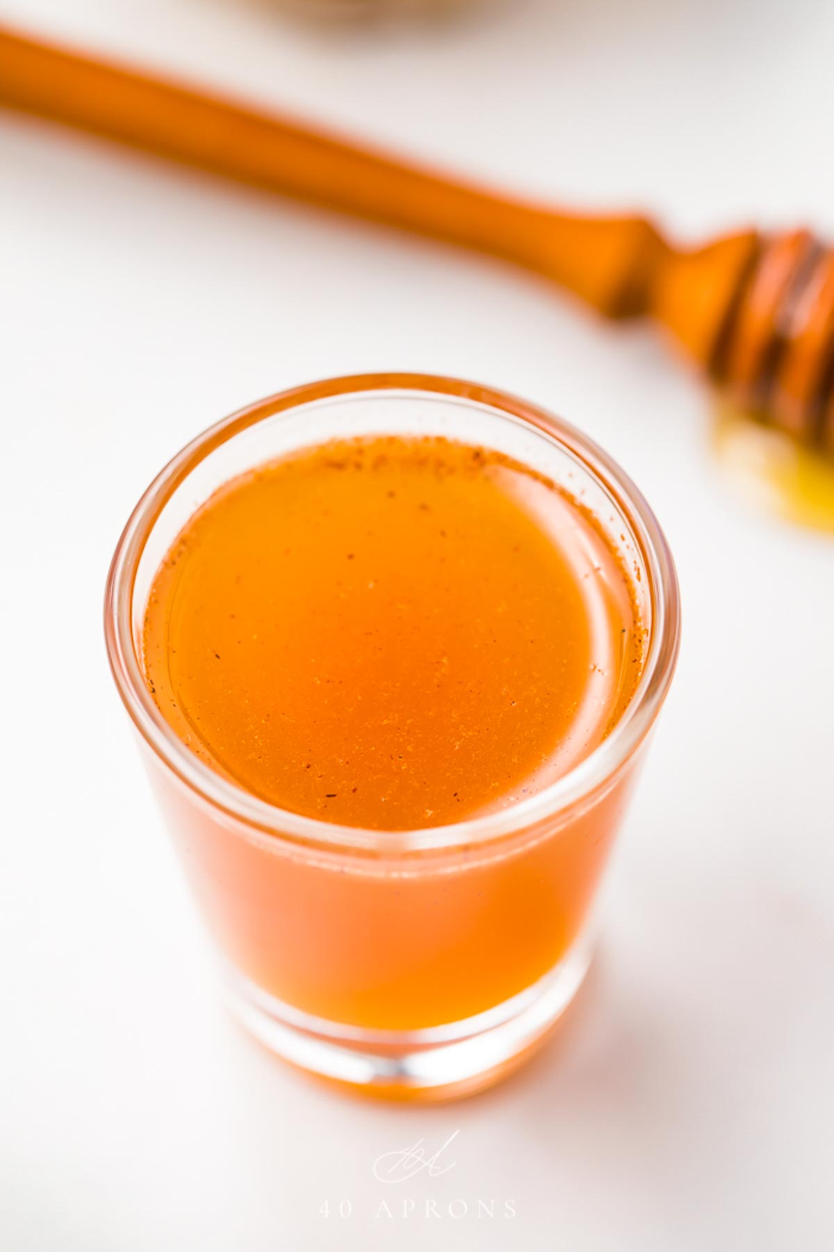 A clear glass shotglass holding an orange liquid peppered with cinnamon, in front of a honey wand dripping with honey.