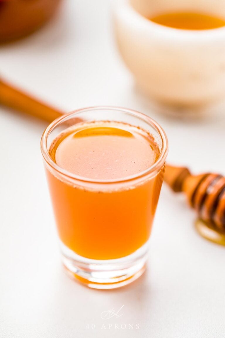 A clear glass shotglass holding an orange liquid peppered with cinnamon, in front of a honey wand dripping with honey.