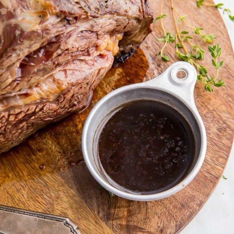 Overhead view of a small silver ramekin holding deep, rich au jus next to a holiday prime rib.