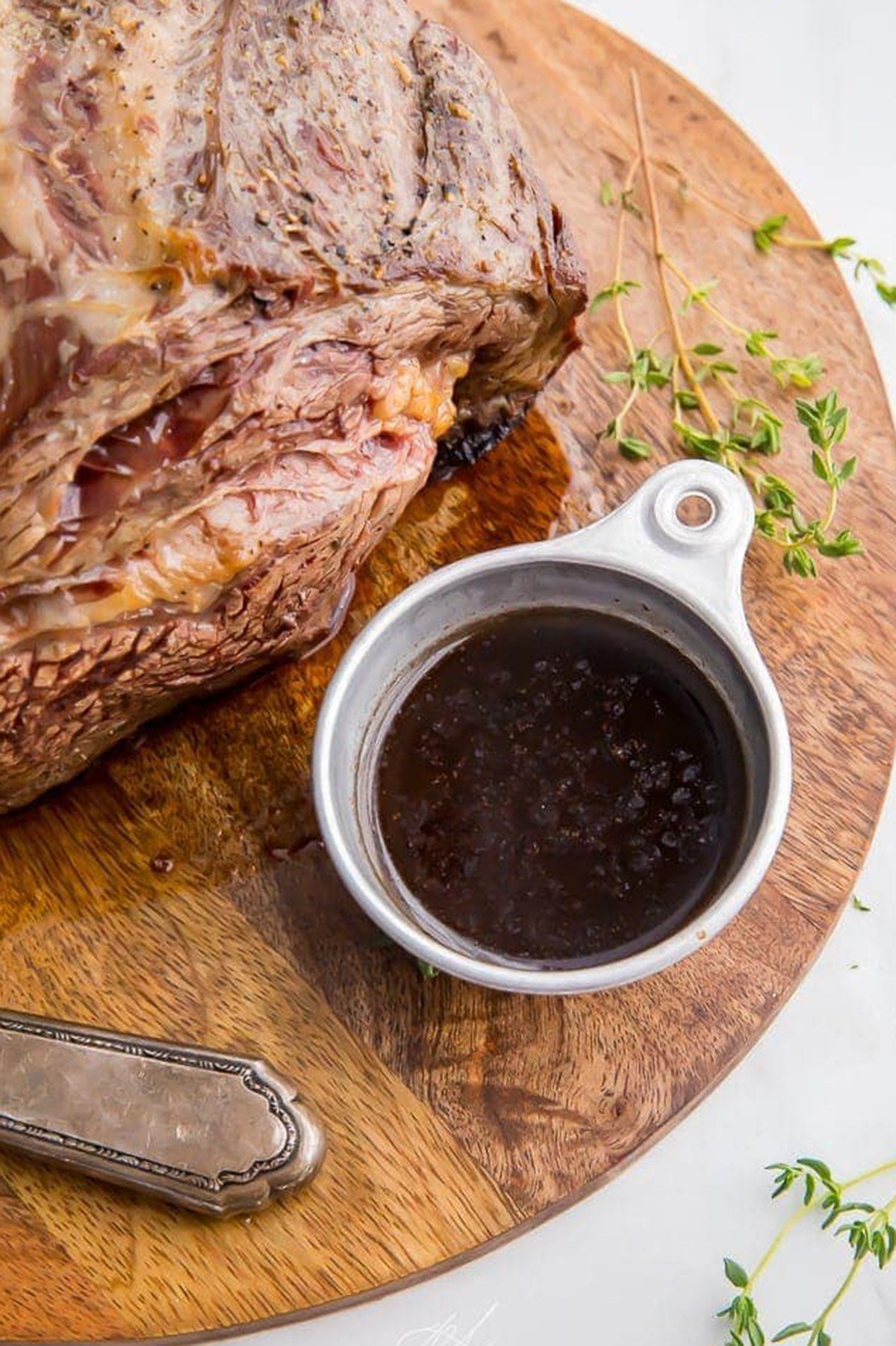 Overhead view of a small silver ramekin holding deep, rich au jus next to a holiday prime rib.