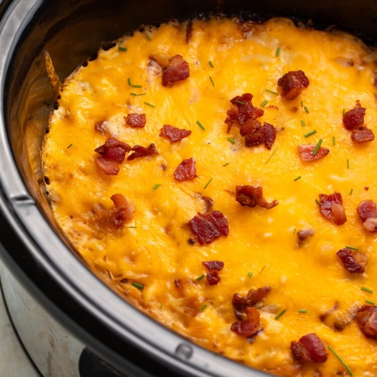 A black Crockpot insert, angled up toward the left of the photo, holding a breakfast casserole topped with melted cheddar and pieces of bacon.