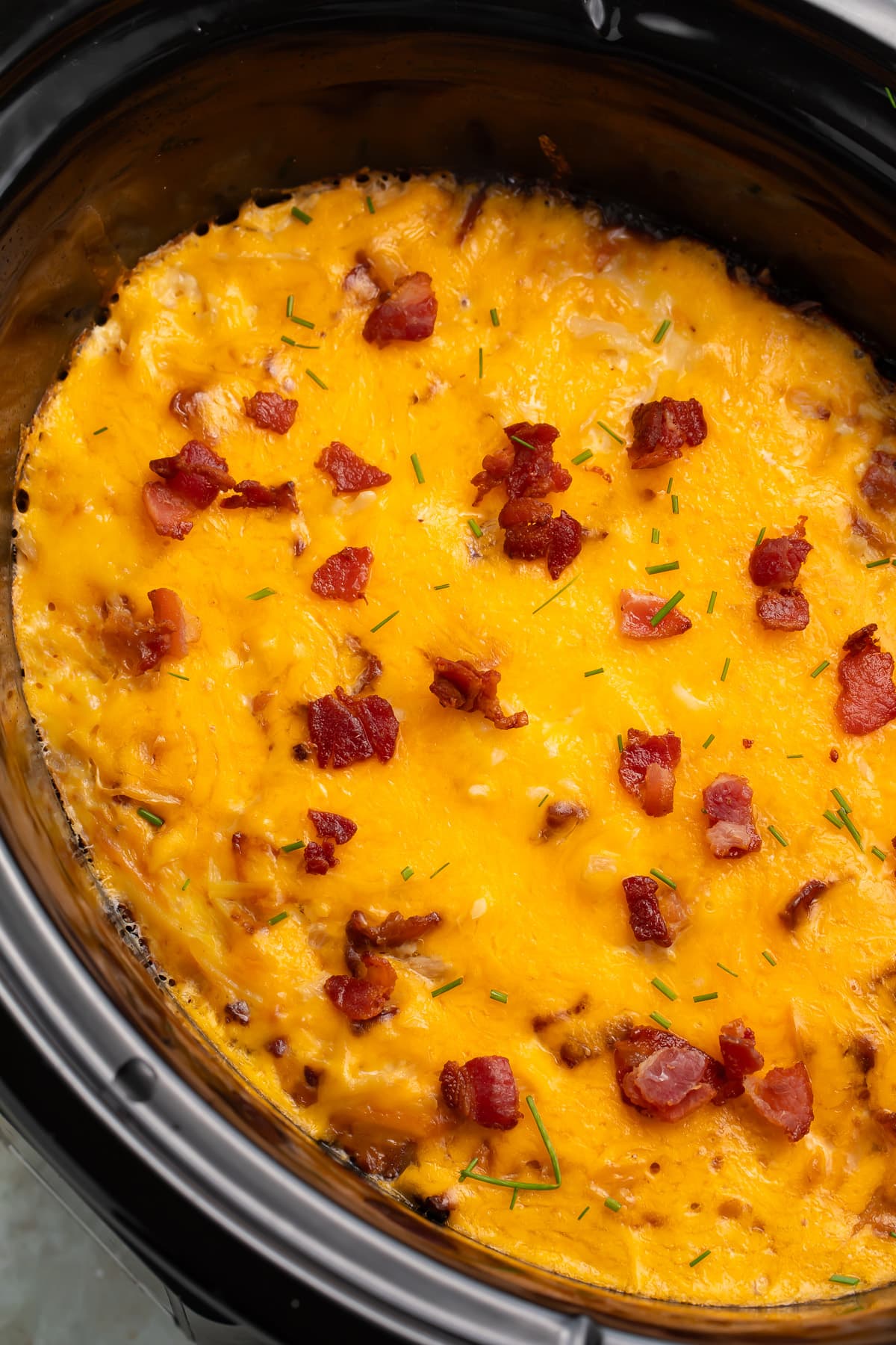 A black Crockpot insert, angled up toward the left of the photo, holding a breakfast casserole topped with melted cheddar and pieces of bacon.