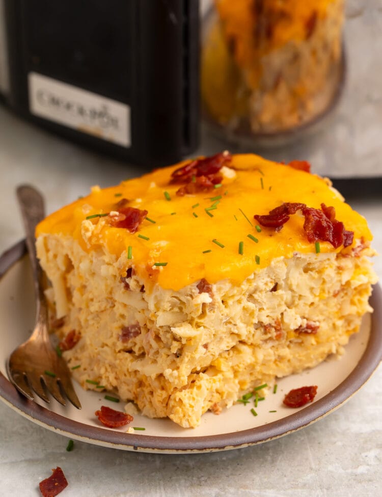 A square, multi-layered slice of Crockpot breakfast casserole on a plate in front of a slow cooker.