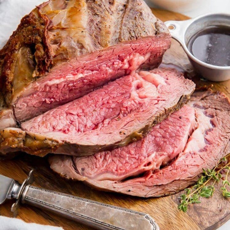 Carved, juicy prime rib resting on a wooden cutting board next to a small silver ramekin of au jus.
