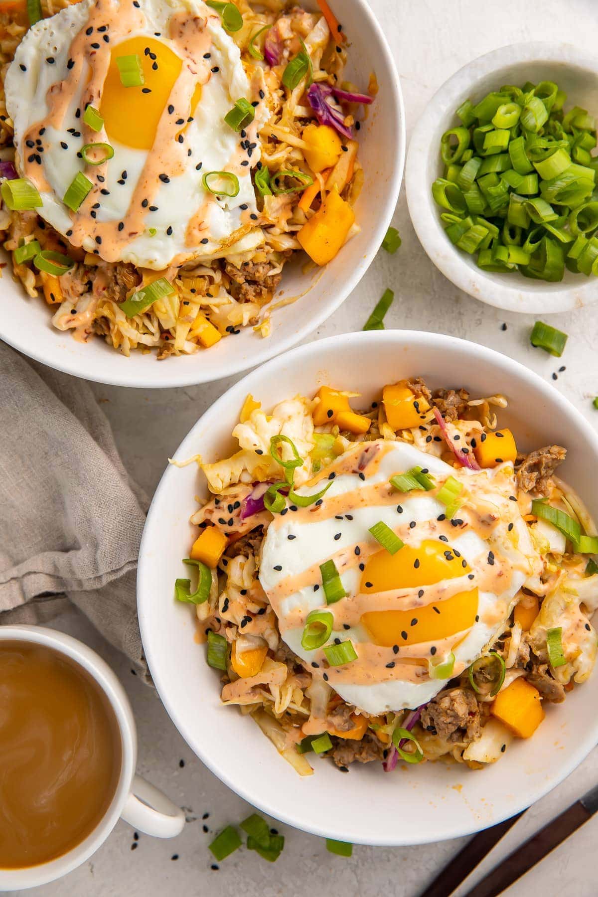 Two white bowls holding a coleslaw, mango, pork sausage mixture topped with fried eggs and green onions