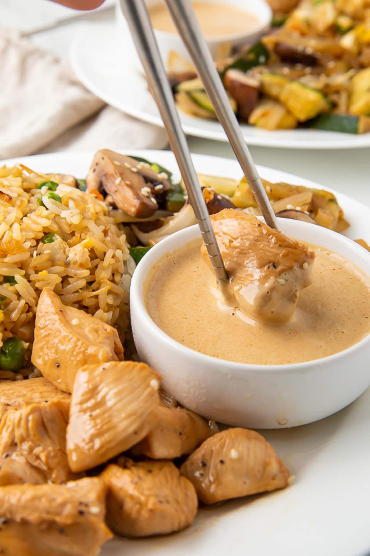 A piece of hibachi chicken being dipped into a creamy mustard sauce on a plate with hibachi fried rice and veggies.