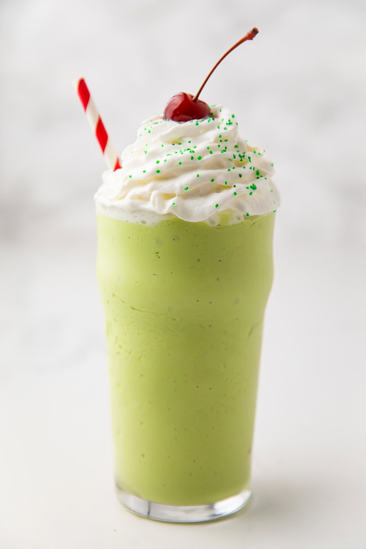 A green dairy-free shamrock shake copycat in a glass topped with whipped cream, a cherry, and a red-and-white striped straw.