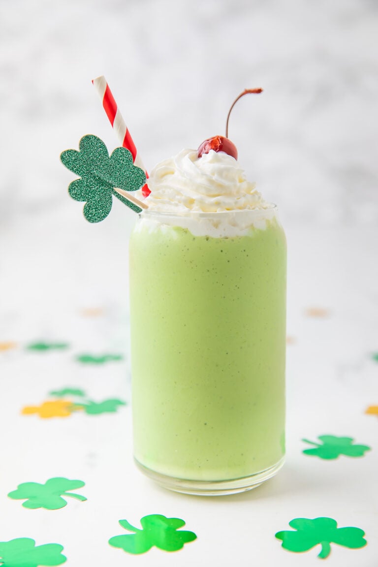 A green dairy-free shamrock shake copycat in a glass topped with whipped cream, a cherry, and a glittery four-leaf clover decoration.