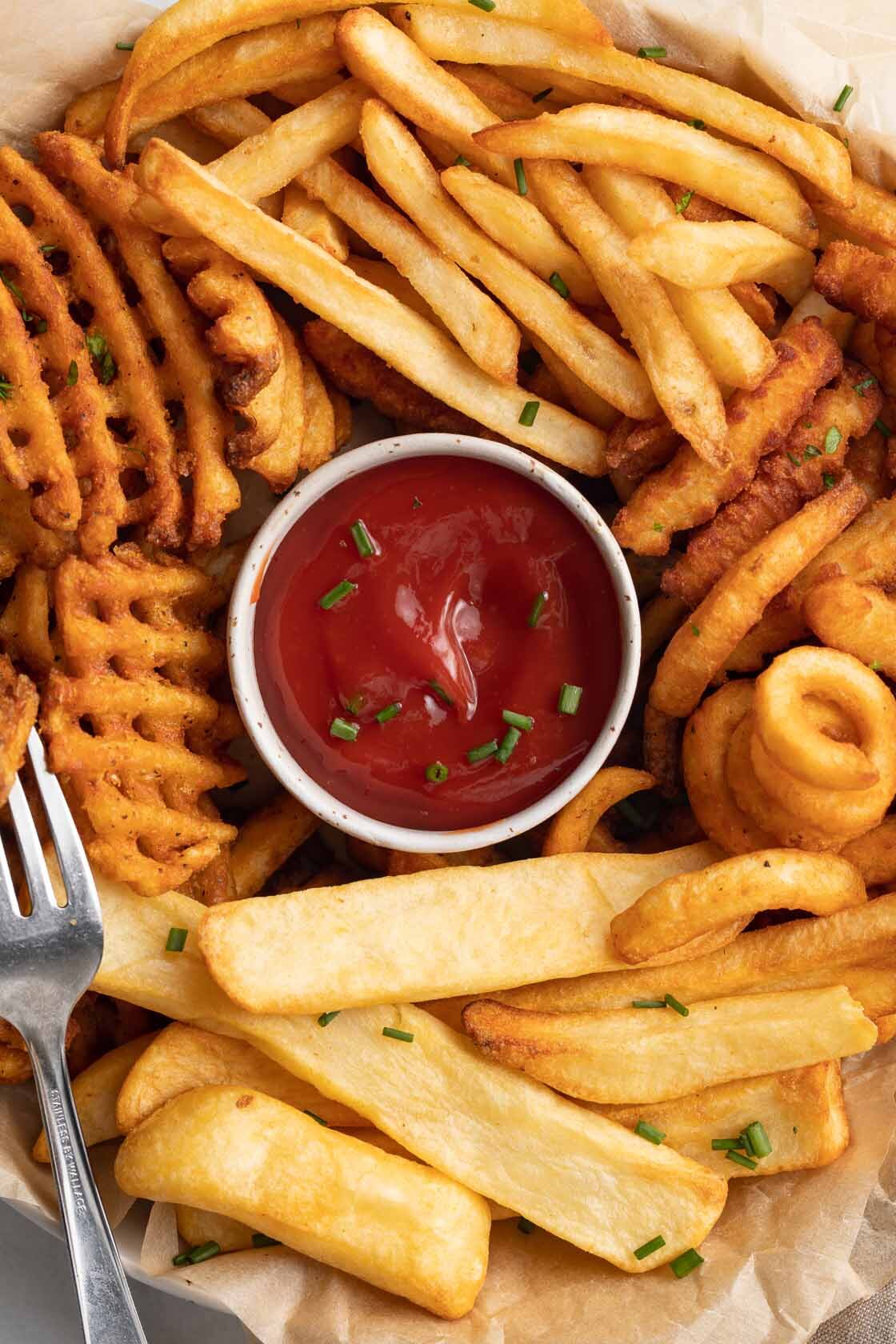 Close-up of different styles of air fryer cooked frozen french fries on a platter, surrounding a ramekin of ketchup.