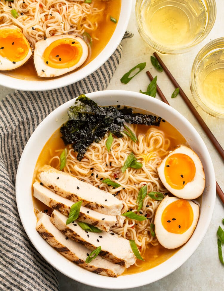 Top-down view of two white bowls holding spicy ramen noodles in broth with slices of chicken, two yolky egg halves, and green garnish.