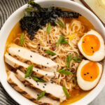 Top-down view of a white bowl holding spicy ramen noodles in broth with slices of chicken, two yolky egg halves, and green garnish.