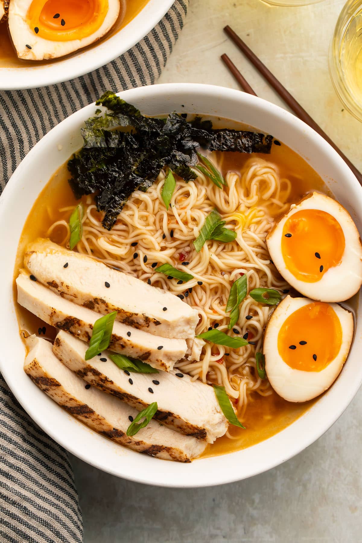 Top-down view of a white bowl holding spicy ramen noodles in broth with slices of chicken, two yolky egg halves, and green garnish.