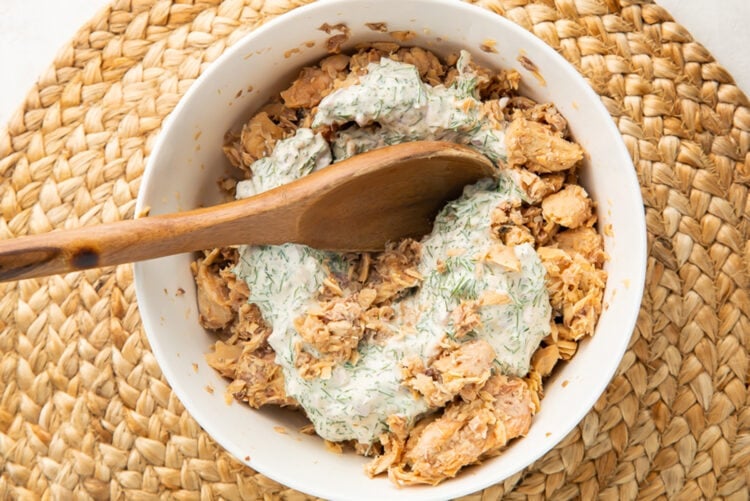 Creamy dill sauce added to a bowl of canned salmon salad with a wooden spoon.