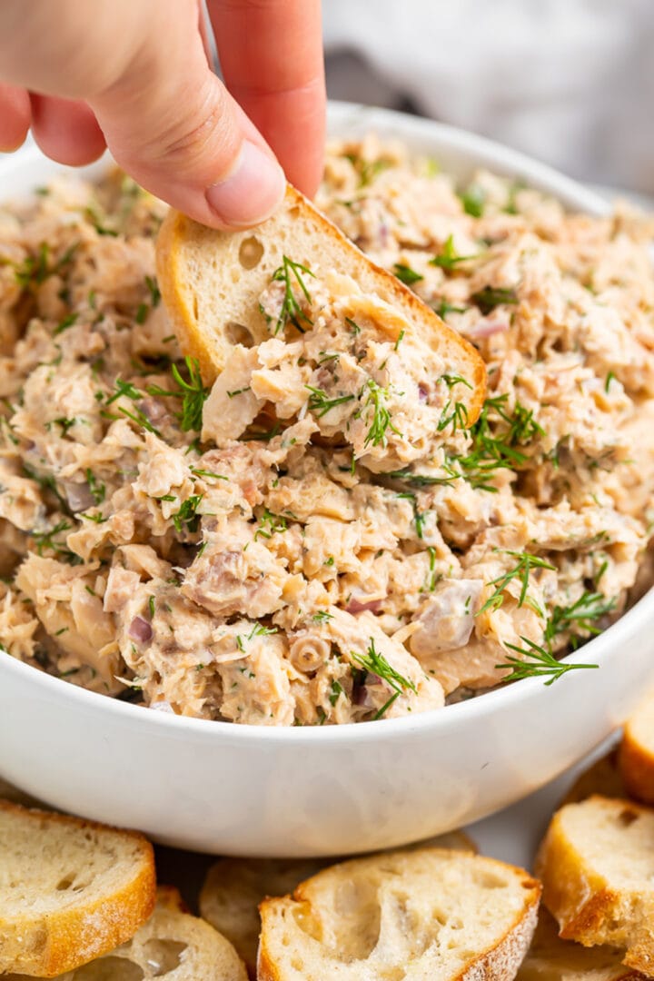 A woman's hand holding a crostini used to scoop salmon salad out of a large bowl.