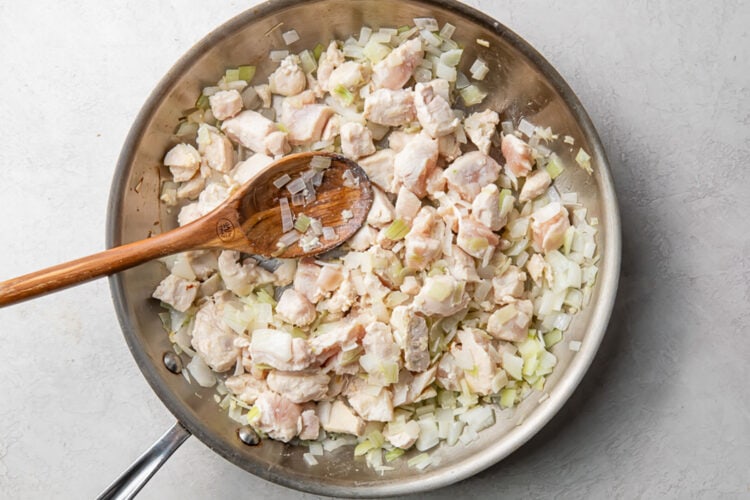 Sautéed onions and garlic with bite-sized pieces of chicken in a large silver skillet with a wooden spoon.