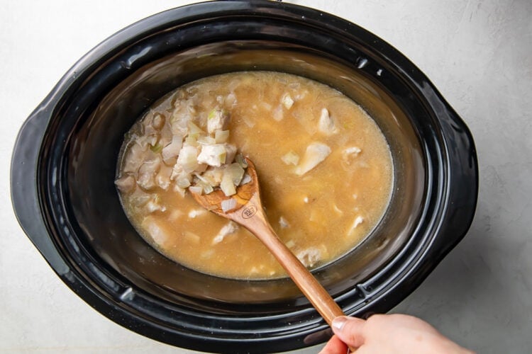 Ingredients for Crockpot chicken and rice in a black Crockpot insert with a wooden spoon.