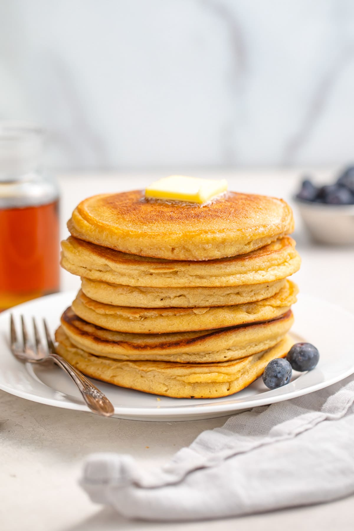 A stack of 6 paleo pancakes resting on a plate next to 2 blueberries. A pat of butter sits atop the pancakes.