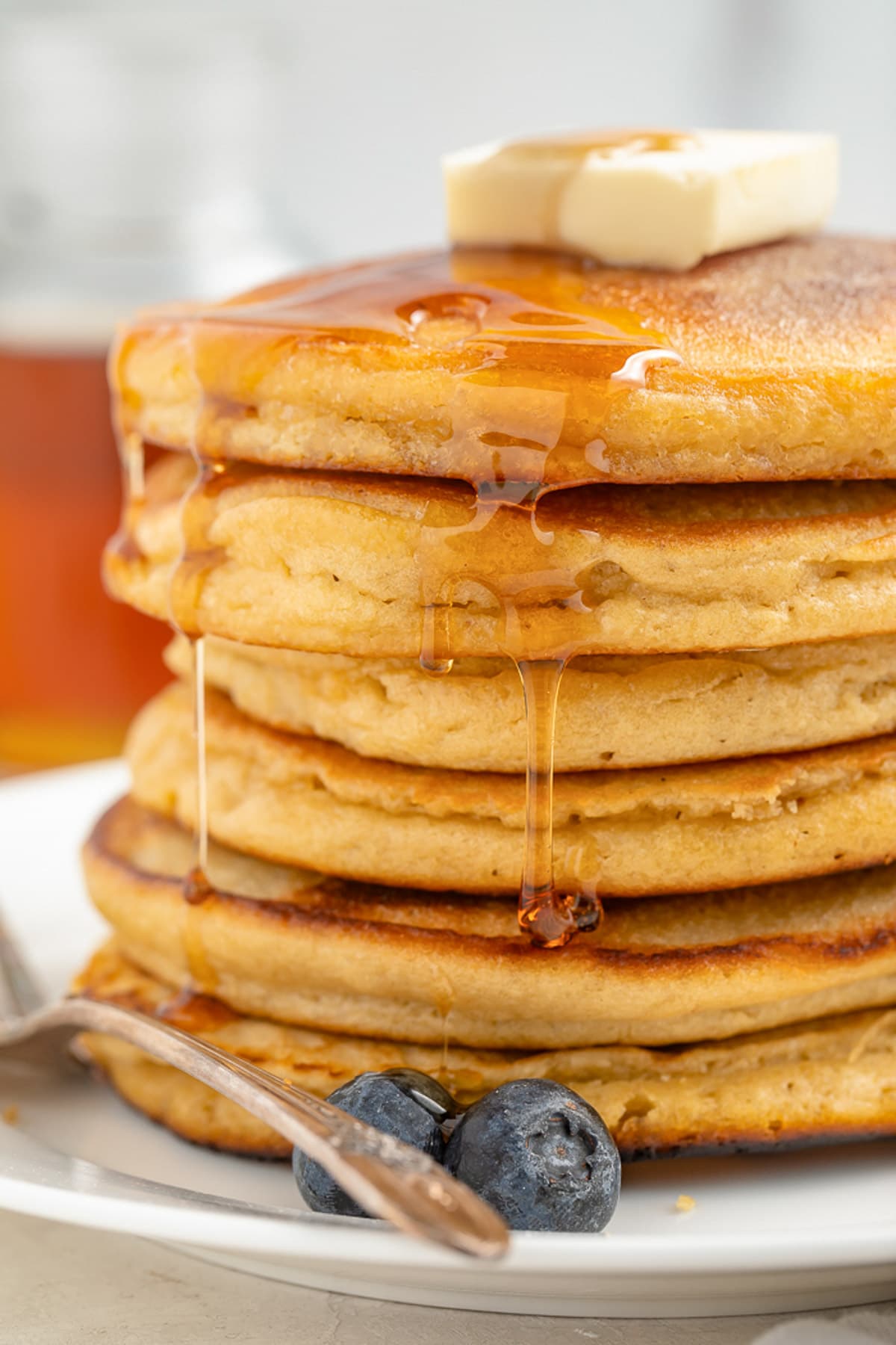 A stack of 6 paleo pancakes on a plate. 2 blueberries rest next to the pancakes, which drip with maple syrup.